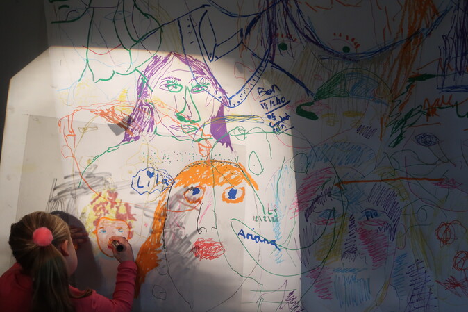 A girl draws on a wall filled with drawings of faces