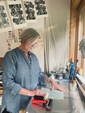 Photo of a woman printmaking. She has short grey hair and is wearing a light blue blouse