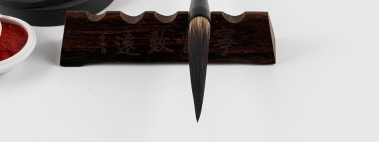 A traditional asian calligraphy brush with a pointed tip, resting on a wooden stand with japanese characters, alongside a red ink container and a black stone on a white background.