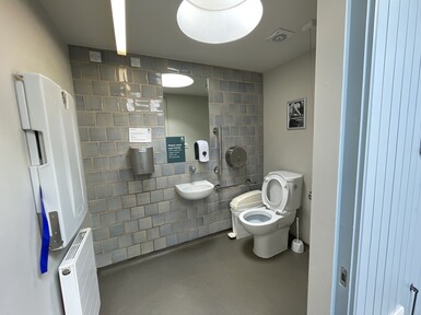 An accessible toilet. The room has a tiled wall, with a toilet and handrails. There is a fold-out changing facility on the wall.