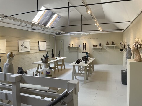 The gallery has off-white walls, there are prints on the wall and sculptures on two tables.