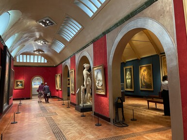 The gallery has red walls and a wooden slatted floor. There is a wall with three arches through which there is another room