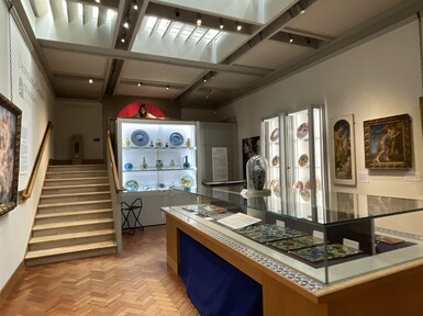 A white room with paintings on the wall, in the middle is a glass case containing decorative tiles