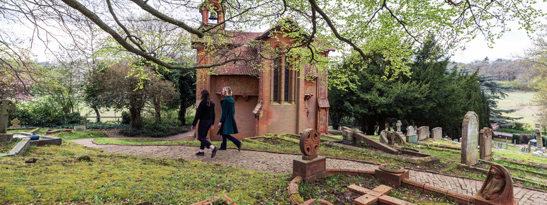 Two visitors walk along the cemetery path, the terracotta chapel is in the background