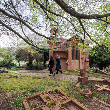 Two visitors walk along the cemetery path, the terracotta chapel is in the background
