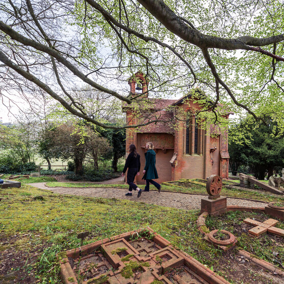 Two visitors walk along the cemetery path, the terracotta chapel is in the background