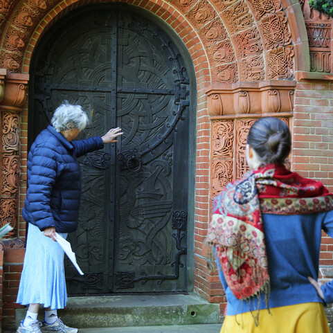 A guide points at the Watts Chapel door, a visitor stands with their back to the camera, looking at the door