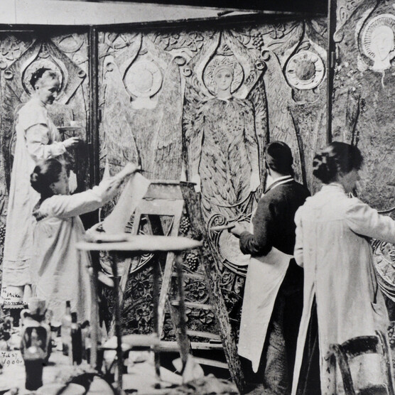 A black and white photograph of Mary with three villagers decorating a highly-decorated panel wall.