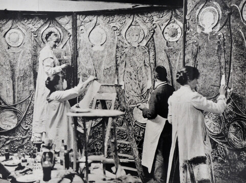 A black and white photograph of Mary with three villagers decorating a highly-decorated panel wall.