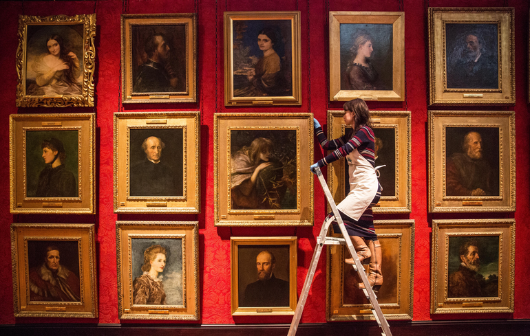 An art technician stands on a ladder high up, inspecting the art works framed on the wall