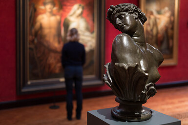 A bronze sculpture of Clytie, a bust turning her head in profile. In the background a visitor views paintings on the red wall.