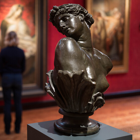 A bronze sculpture of Clytie, a bust turning her head in profile. In the background a visitor views paintings on the red wall.