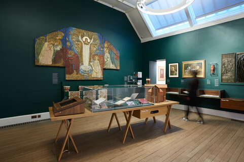 A green gallery with artworks on the wall and a wooden table with a glass case, filled with objects