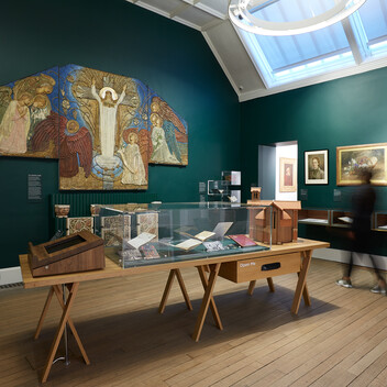 A green gallery with artworks on the wall and a wooden table with a glass case, filled with objects
