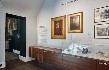 A white gallery with a glass display case and paintings on the wall