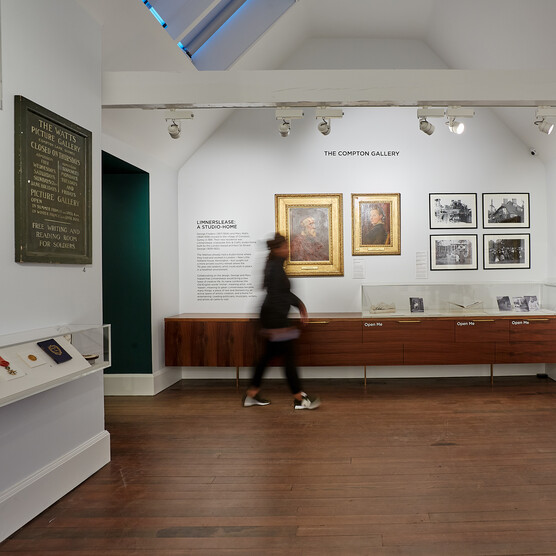 A visitor walks in the white-walled gallery. On the wall are paintings and photos.
