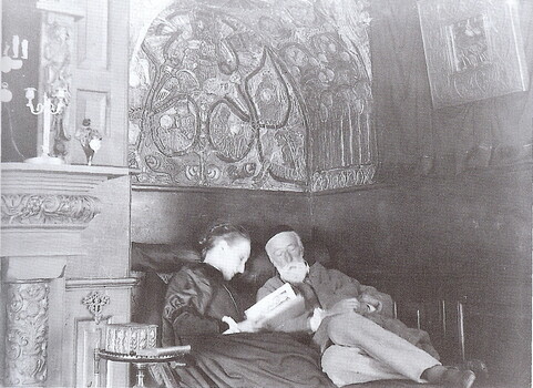 Archive black and white photo of George and Mary, Mary reads to George underneath a decorated arch way