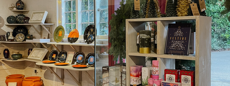 Photograph of the inside of a shop. There are various Christmas decorations displayed on the table in the foreground, such as terracotta Christmas tree decorations and festive candles. there are some large terracotta plant pots placed on the floor an on the shelves in the background.