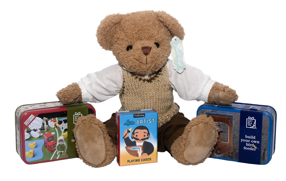 Product photograph of shop items. In the middle of the image is a brown teddy bear with a white shirt and beige knitted vest on, and in front of the bear is a kids card game with a cartoon of Salvadore Dali on the front. To the left of the bear is a red tin with a toy farm scene on the front, and to the right of the bear is a blue tin with a photo of a bird feeder on the front.
