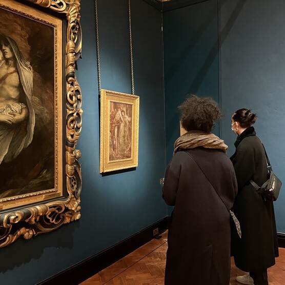 Two visitors with their backs to camera view a gold-framed painting in the blue-walled Historic Gallery