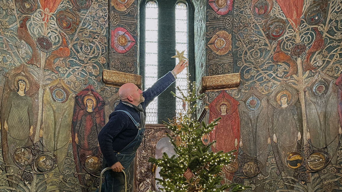Photograph of a man standing on a ladder and placing a star on the top of a Christmas tree inside Watts Chapel. The walls of the chapel are decorated with angels made from painted gesso.