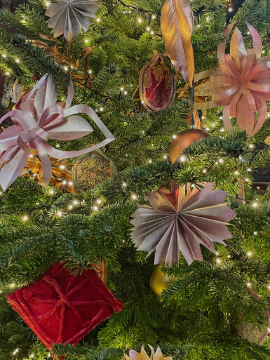 Close-up photograph of a Christmas tree. Decorating the tree are fairy lights, hand painted terracotta decorations and other decorations made out of paper and card.
