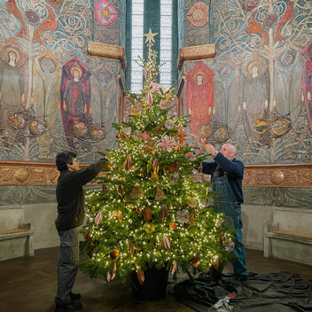 Photograph of a Christmas tree in the middle of Watts Cemetery Chapel. The walls of the chapel are decorated with angels made from painted gesso. A male and a female are decorating the tree.
